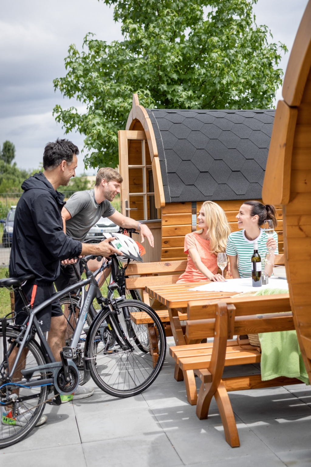 Zwei Frauen sitzen am Tisch zwischen zwei Schlaffässern am Campingplatz der Parktherme Bad Radkersburg. Am Tisch sind auch Weingläser und eine Weinflasche zu sehen. Zwei Männer stehen mit ihren Fahrrädern neben dem Tisch und unterhalten sich mit den Frauen.