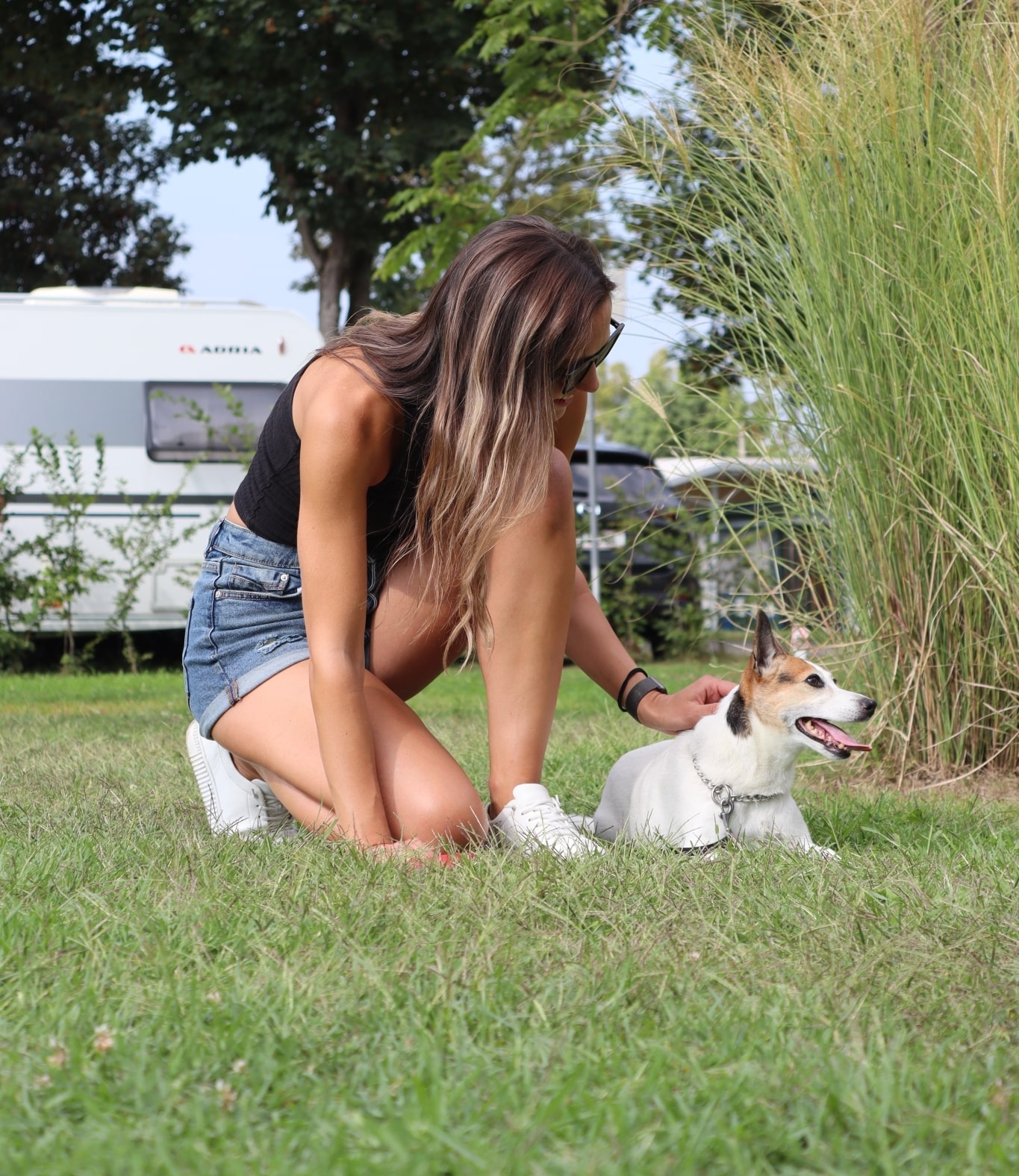 Dame kniet in der Wiese und streichelt einen Hund. Im Hintergrund sieht man einen Campingwagen.