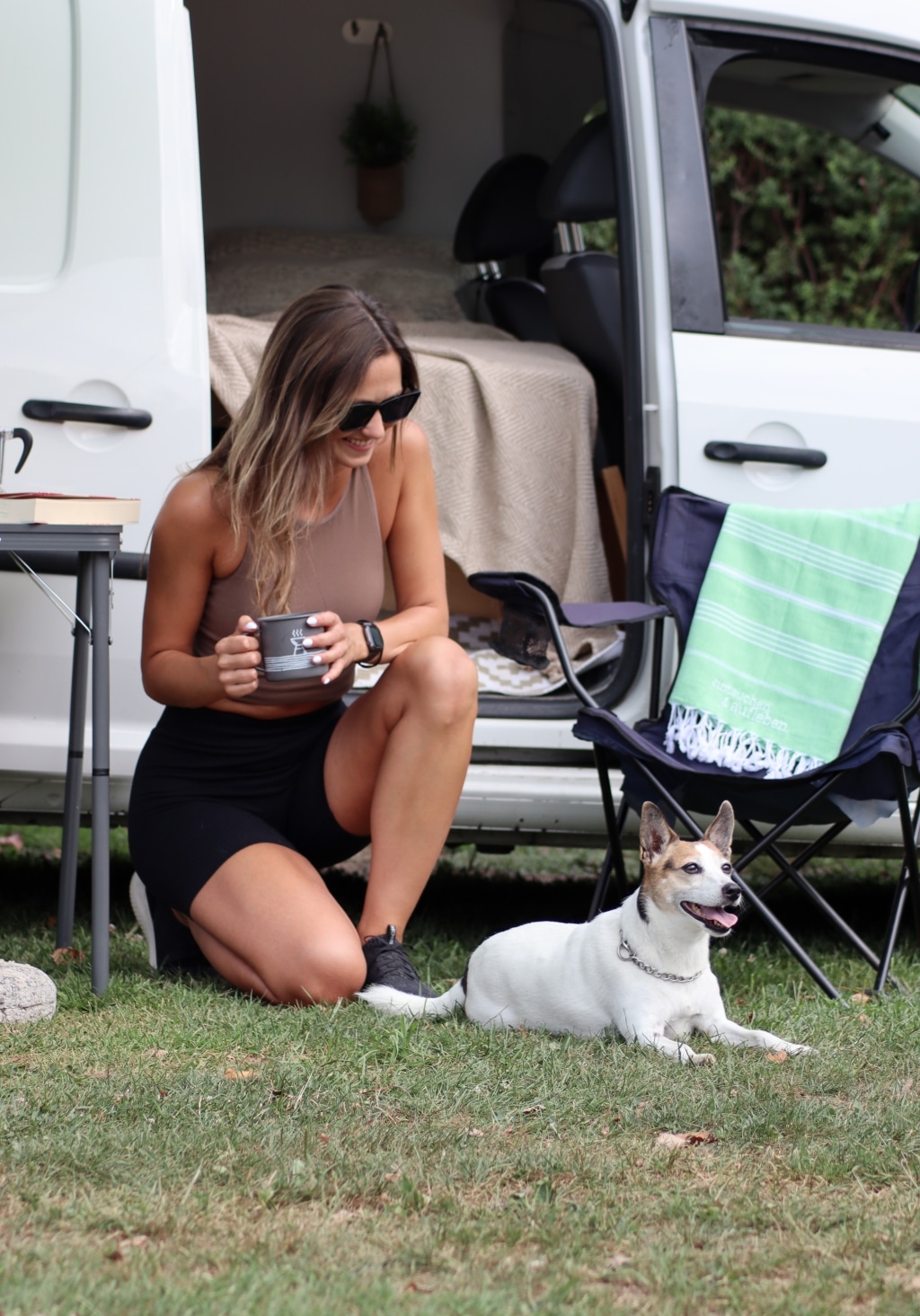 Dame sitzt mit Hund vor einem offenen Campingvan.